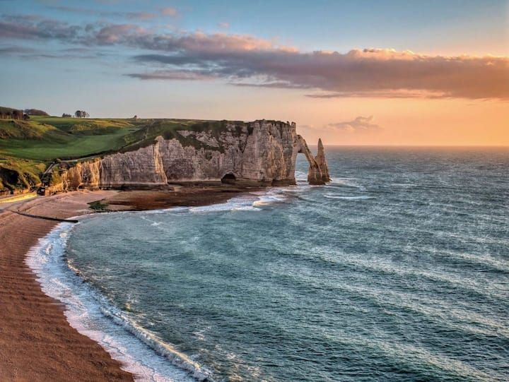 Falaises côtières au coucher du soleil avec arche et plage ; océan aux eaux bleu-vert et ciel rose pâle.