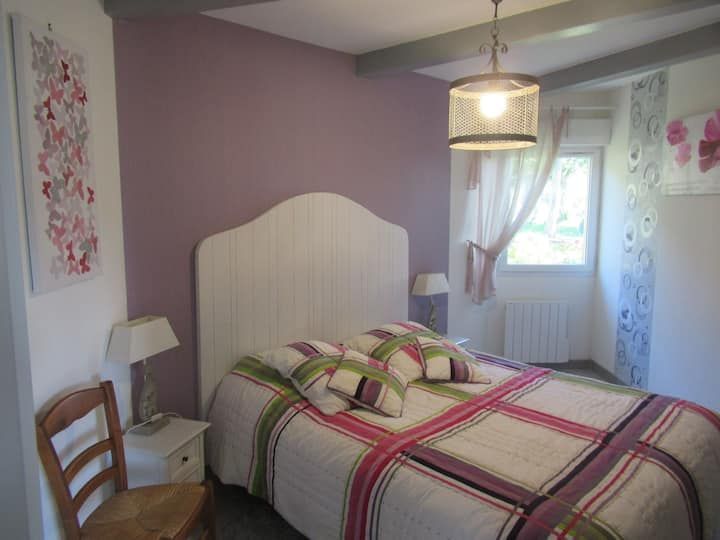 Chambre avec tête de lit blanche, mur violet et linge de lit à motifs floraux. Elle comprend une fenêtre, des œuvres d'art et une suspension lumineuse.