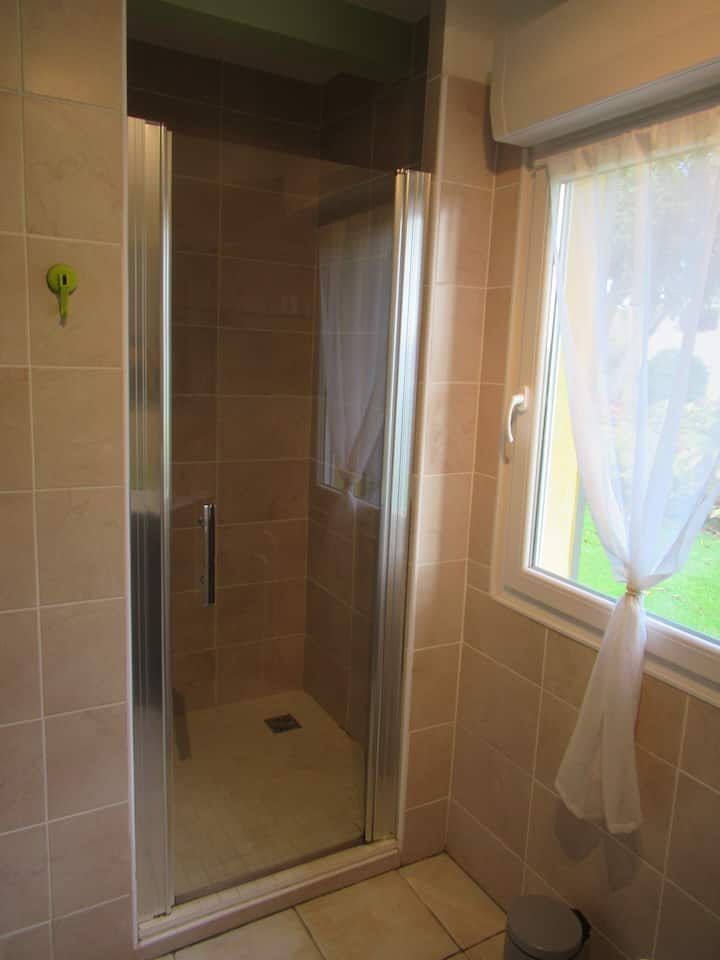 Cabine de douche avec porte en verre, murs carrelés beiges, fenêtre avec rideau blanc et sol beige.