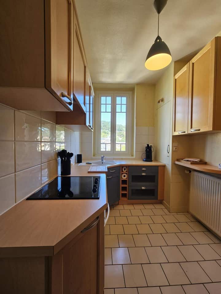 Cuisine avec placards en bois, plans de travail clairs et sol carrelé ; fenêtre au loin.