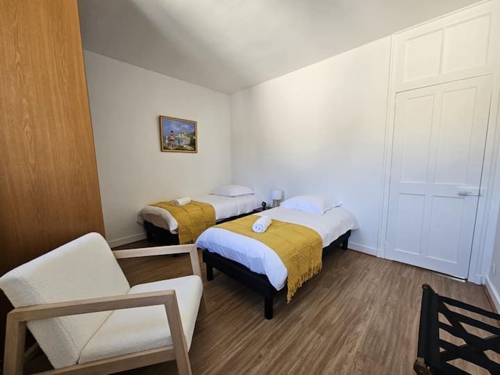 Chambre avec deux lits jumeaux, couvertures moutarde, murs blancs, parquet et un fauteuil.