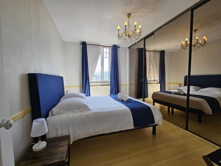 Chambre avec des touches de bleu, notamment une tête de lit, des rideaux et un couvre-lit décoratif. Placard à portes miroir et lustre.
