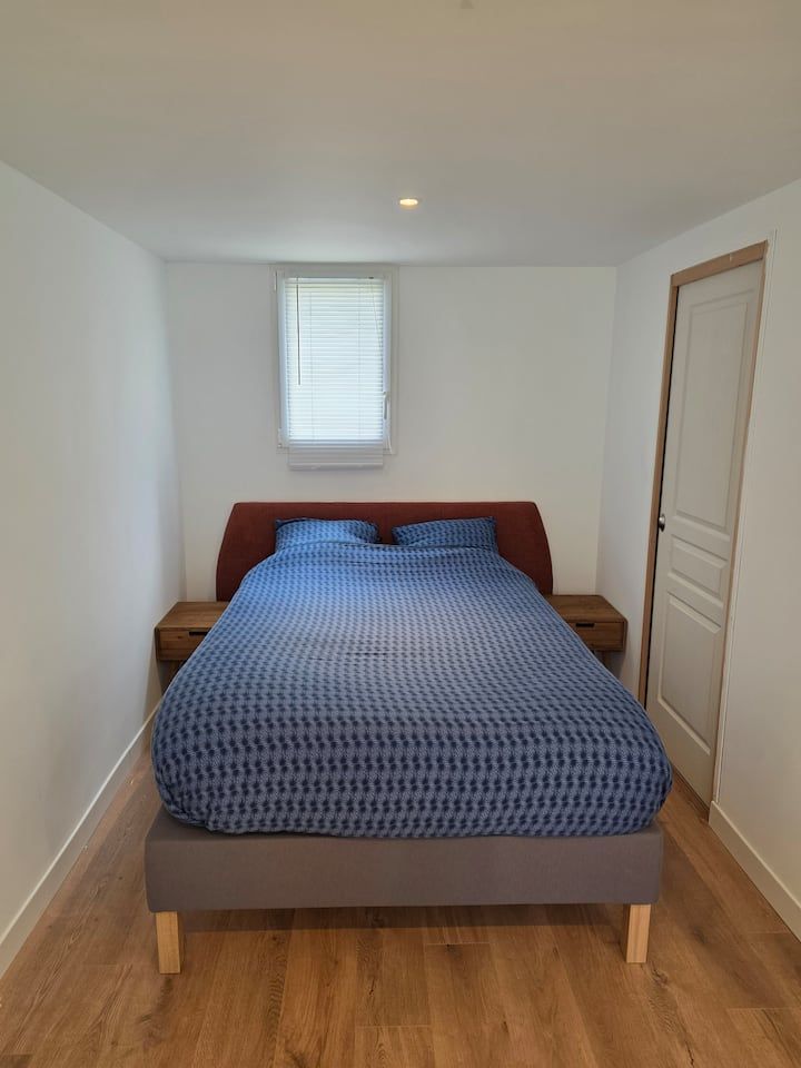 Petite chambre avec un couvre-lit bleu, un parquet, des murs blancs et une fenêtre au-dessus du lit.