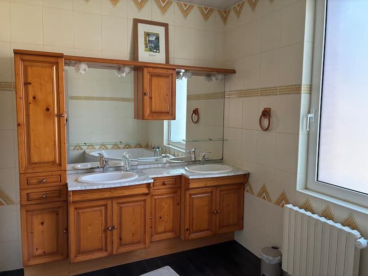 Meuble-lavabo en bois avec lavabo et miroir, murs carrelés, fenêtre et radiateur à côté.