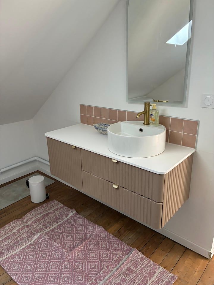 Petit meuble-lavabo avec vasque blanche, robinet doré, miroir et tapis rose sous un plafond mansardé