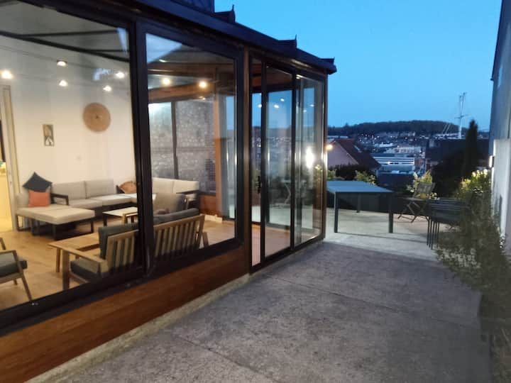 Terrasse vitrée donnant sur la ville au crépuscule, avec sièges extérieurs et éclairage intérieur chaleureux
