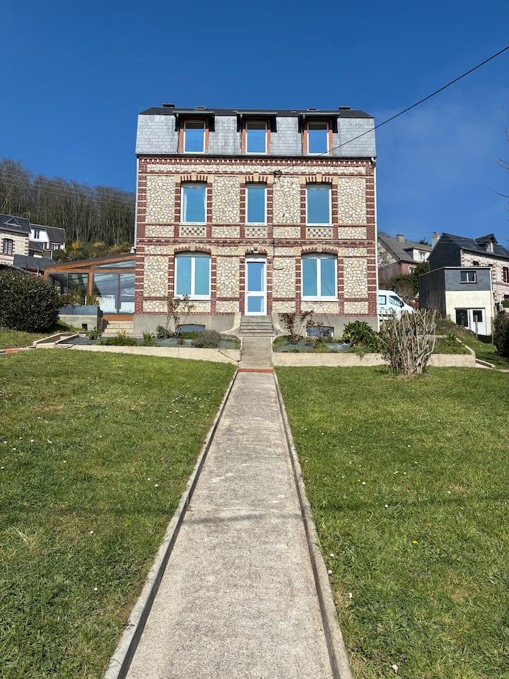 Maison symétrique en briques avec volets bleus, allée centrale et pelouse verte sous un ciel bleu clair