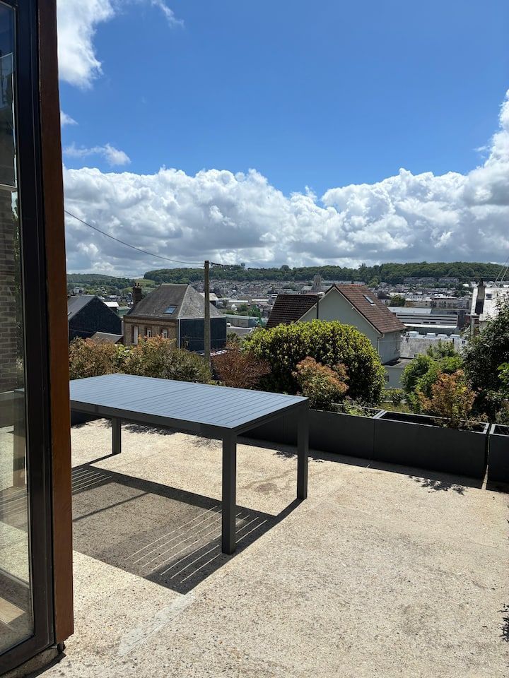Terrasse avec table noire donnant sur une ville ensoleillée et un ciel bleu nuageux