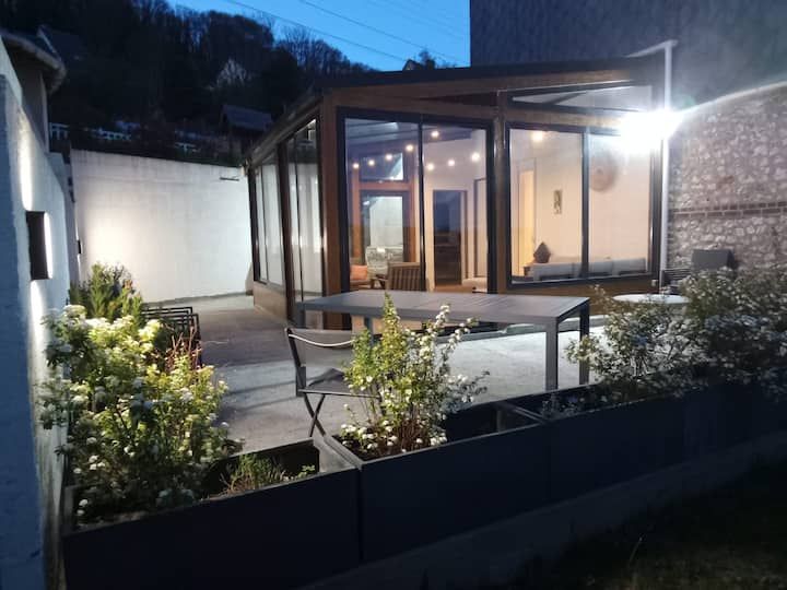 Patio d'une maison moderne au crépuscule, avec éclairage chaleureux, murs de verre, marches et jardin éclairé à l'avant.