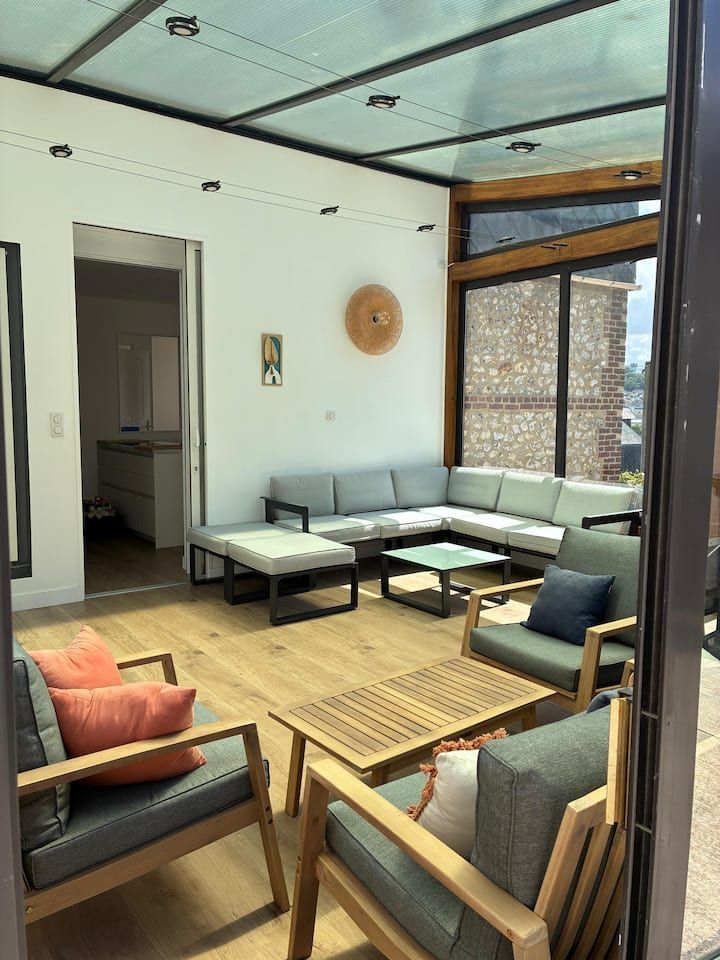 Salon moderne avec meubles en bois clair, canapé gris, fauteuil orange et grande fenêtre.