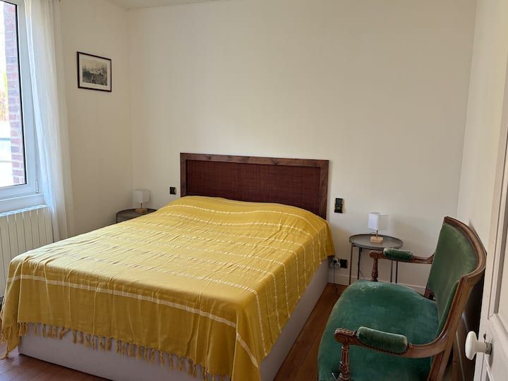 Chambre avec un couvre-lit jaune, une chaise verte, une table de chevet et une fenêtre avec des rideaux blancs.