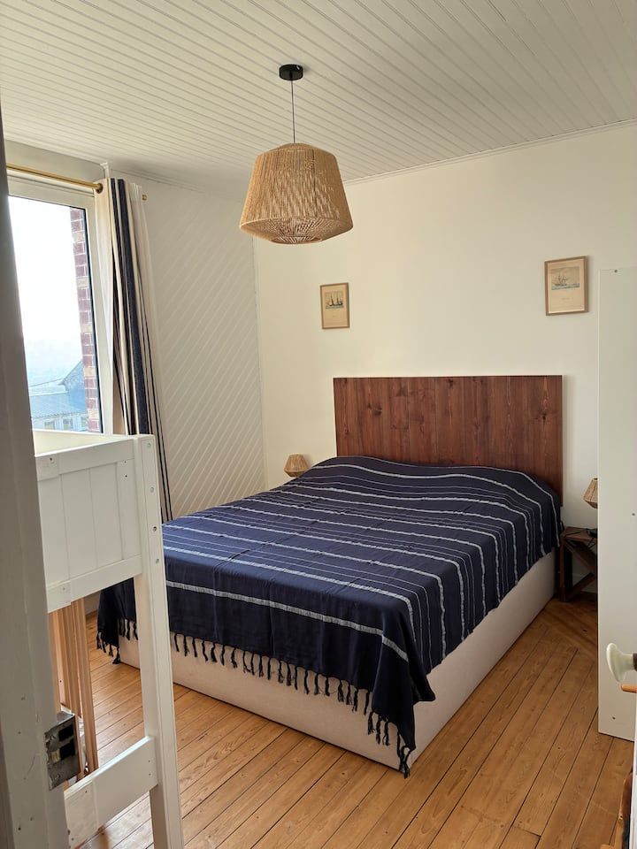 Petite chambre avec un lit en bois, une couverture à rayures bleu marine, une suspension lumineuse en tissu tressé et une fenêtre ouverte.