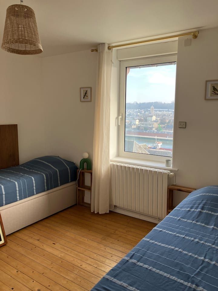 Petite chambre avec deux lits simples, parquet et fenêtre donnant sur un paysage urbain.