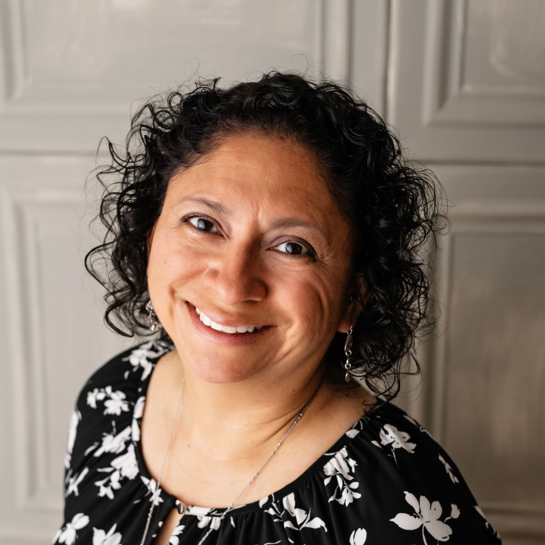 Smiling woman with curly dark hair in a black floral top against a light paneled wall