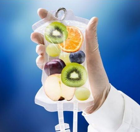 Hand holding IV bag filled with fruit: kiwi, orange, grapes, apple. Blue background.