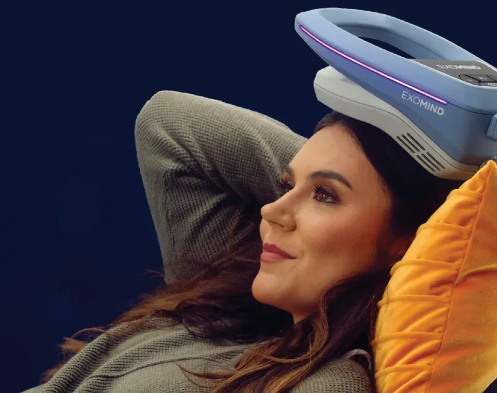 Woman reclining with a device on her head, smiling. Blue and white device. Orange pillow. Dark blue background.