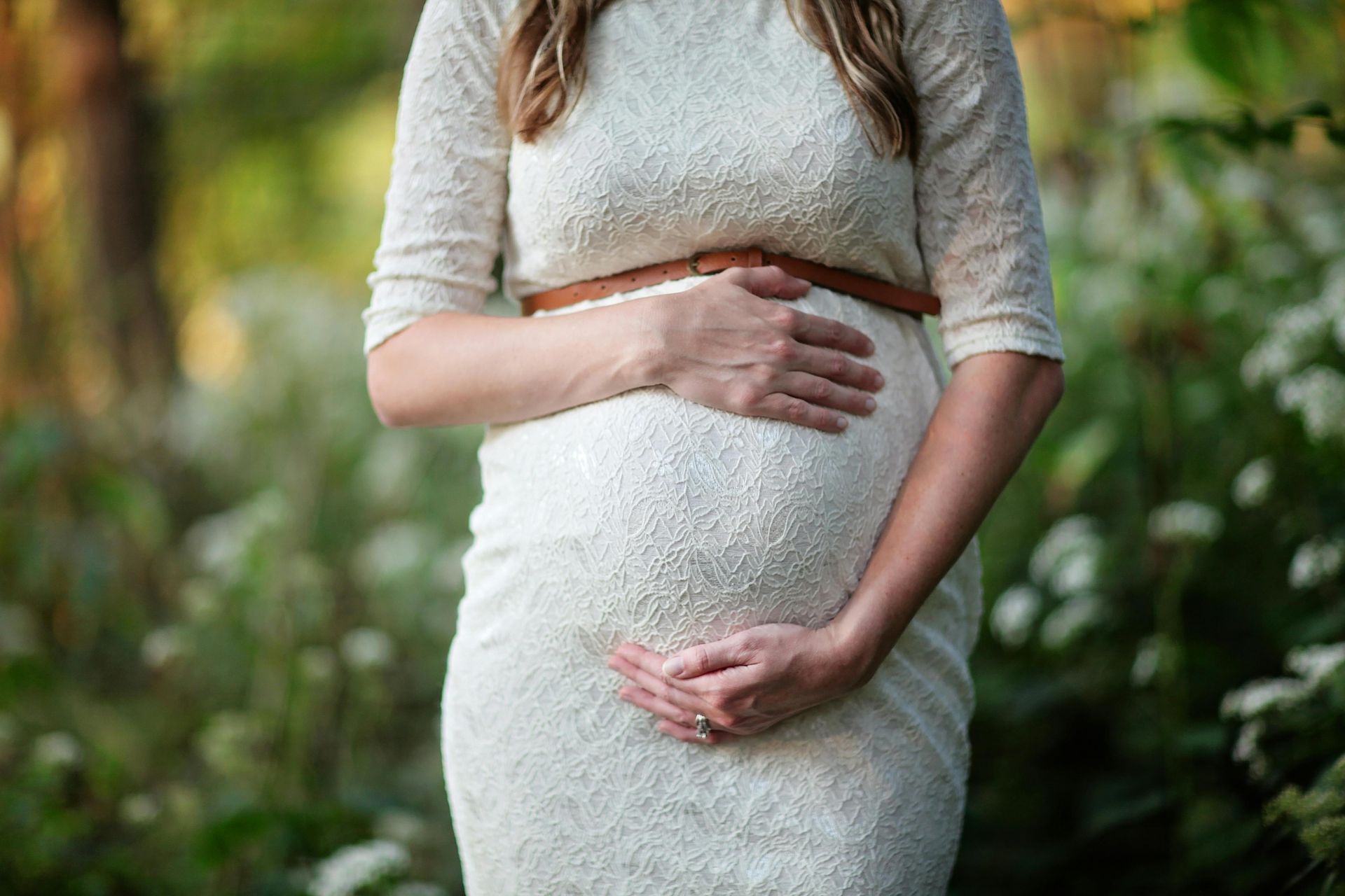 Pregnant person in white lace dress, hands cradling belly, standing outdoors with foliage.