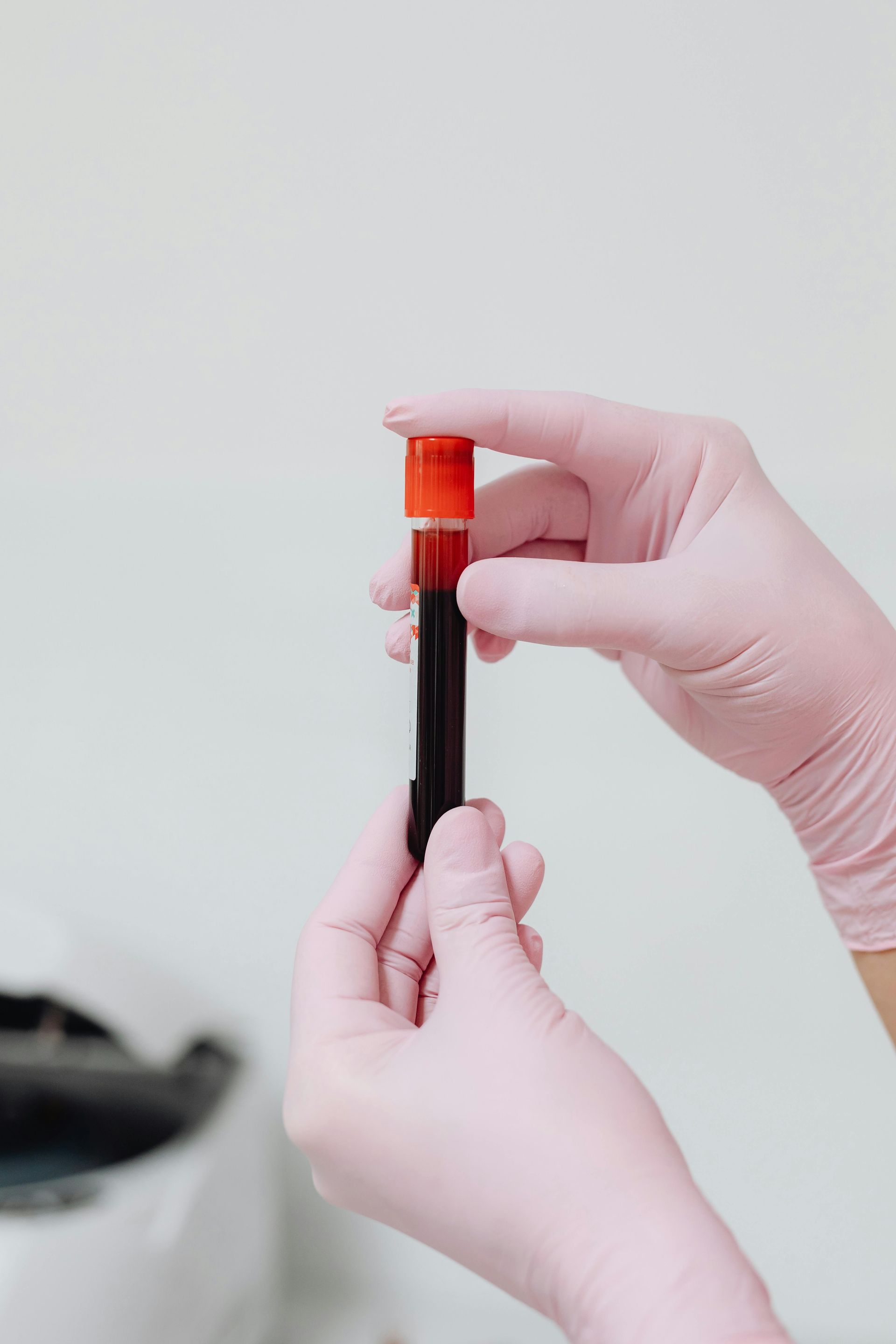 Hands in pink gloves holding a blood sample vial with a red cap.