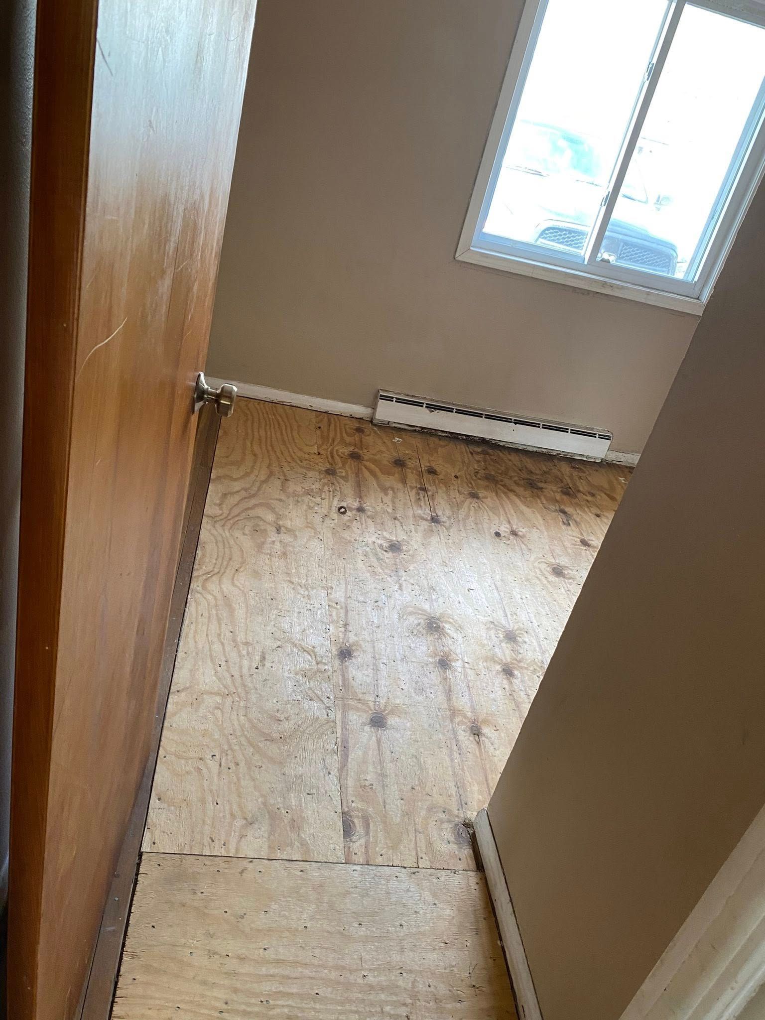 Narrow hallway with unfinished plywood floor and a window at the end.