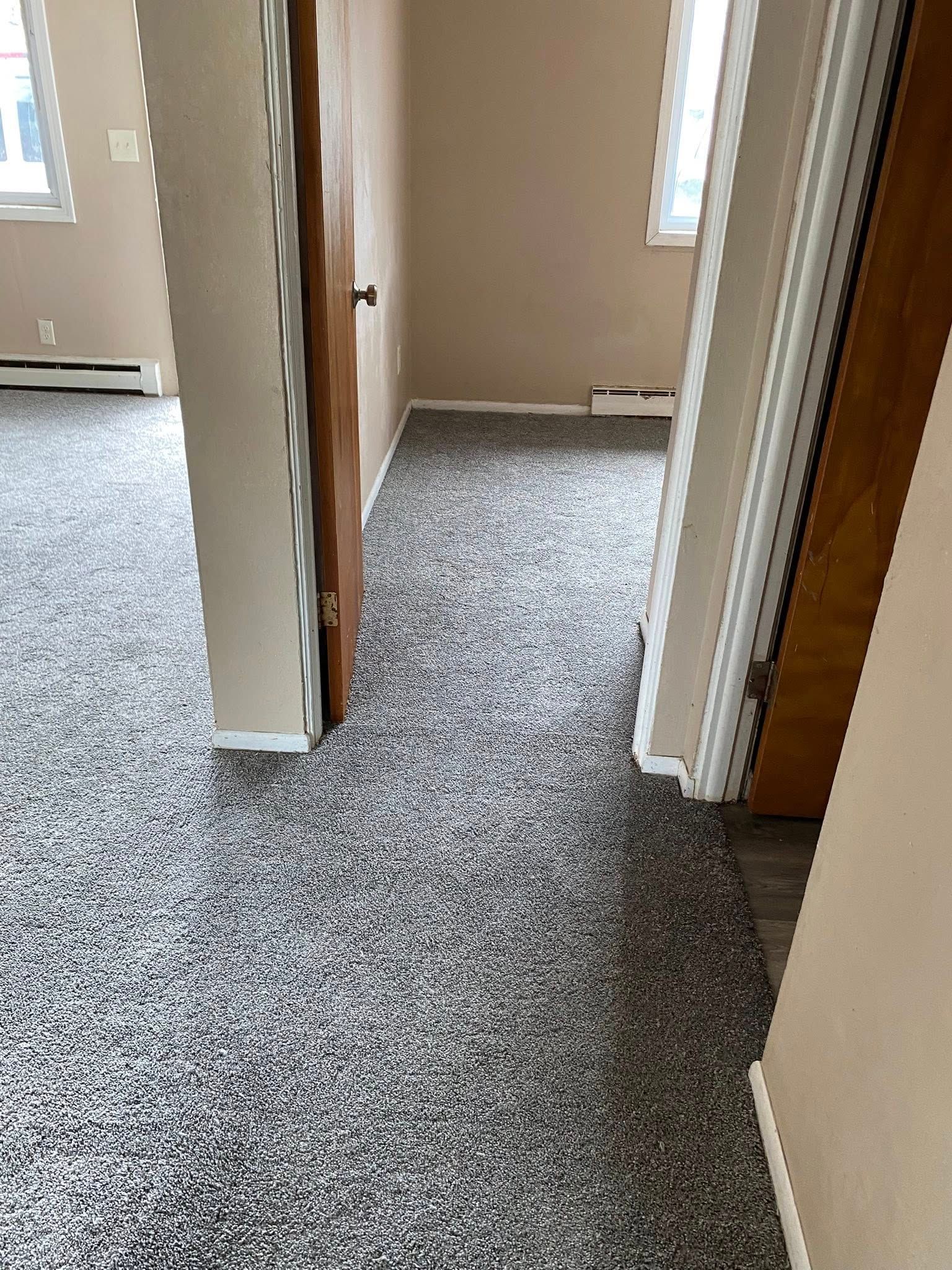 Empty hallway with beige walls, wood trim, and gray speckled carpet, viewed through open doorways