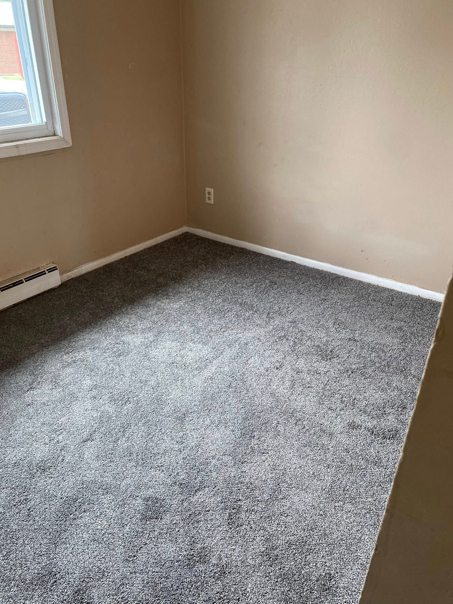 Empty beige room with gray carpet and a small window on the left.