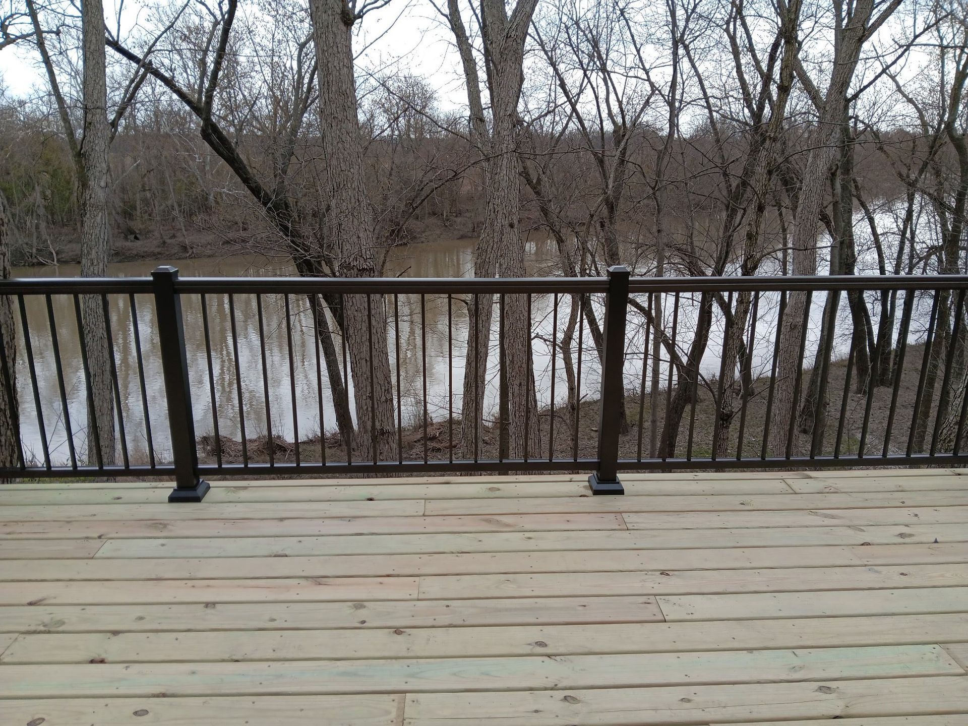 Wooden deck with black railing overlooking a winter river and leafless trees