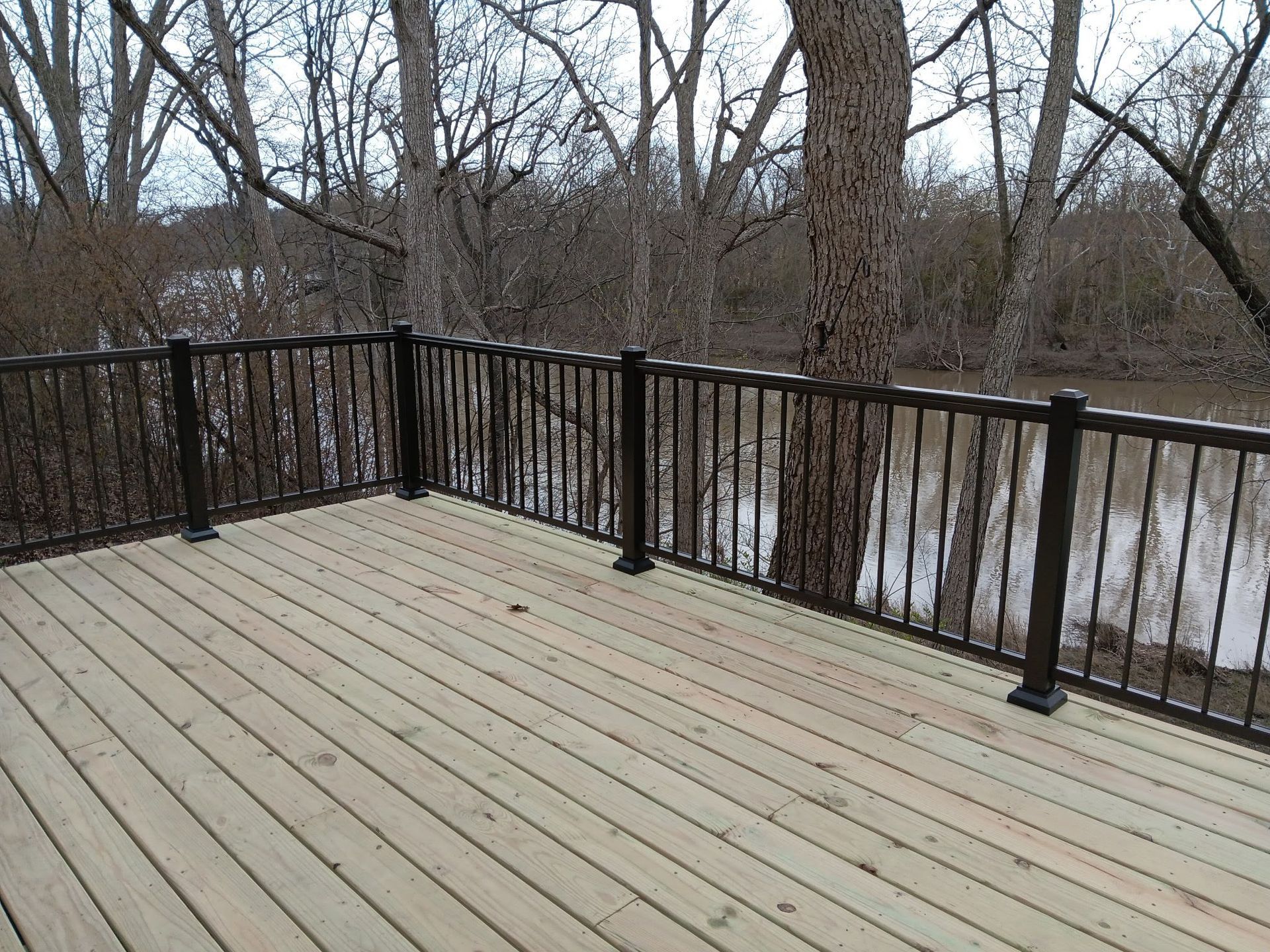 Wooden deck with black metal railing overlooking a wooded, leafless backyard