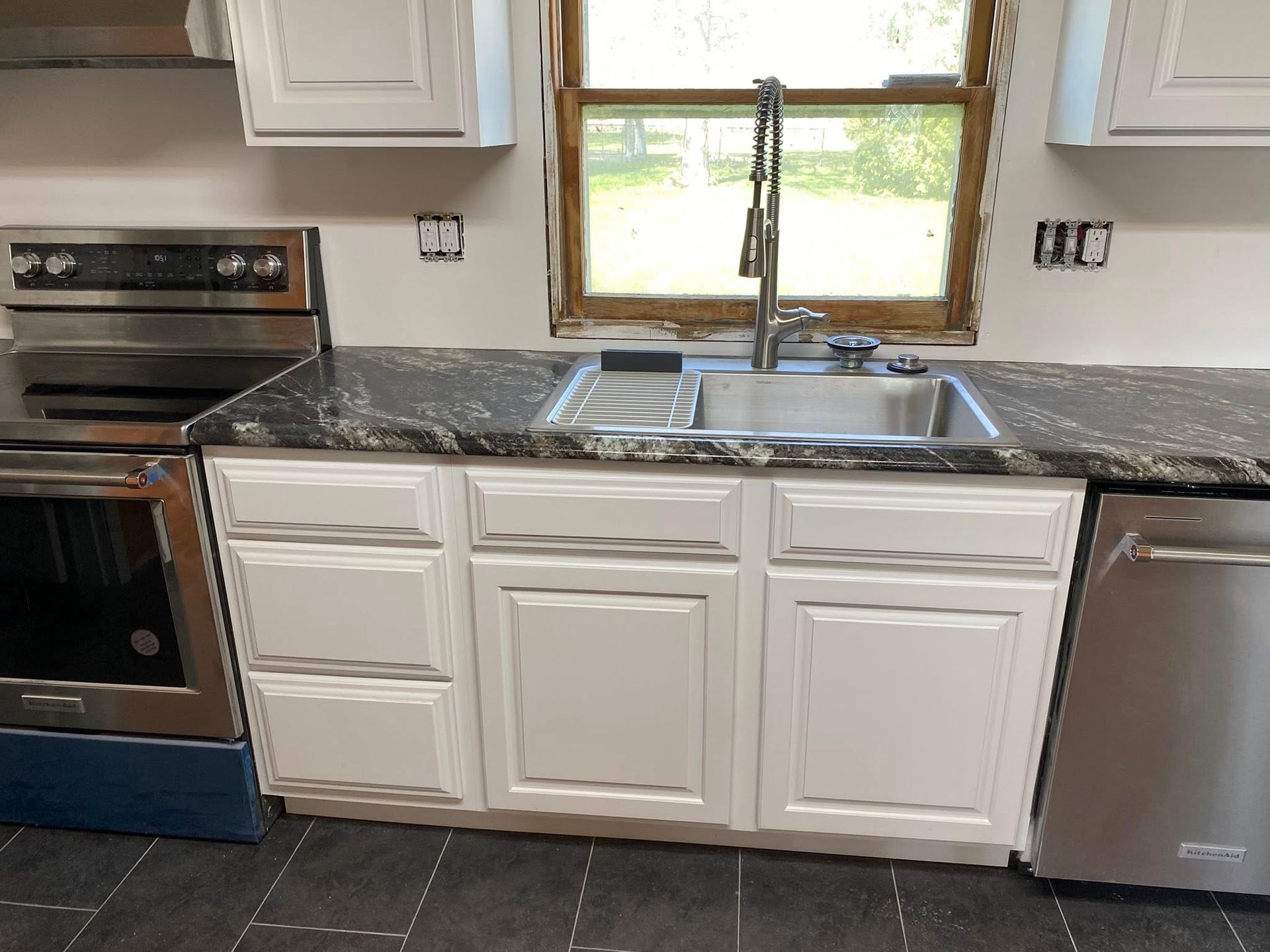 Kitchen sink beneath window, white cabinets, dark countertop, stainless oven and dishwasher.