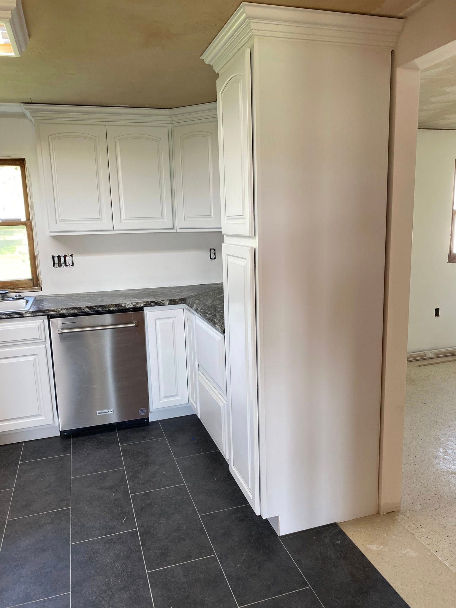 Small kitchen with white cabinets, stainless dishwasher, dark countertop, and a tall pantry cabinet by an archway.