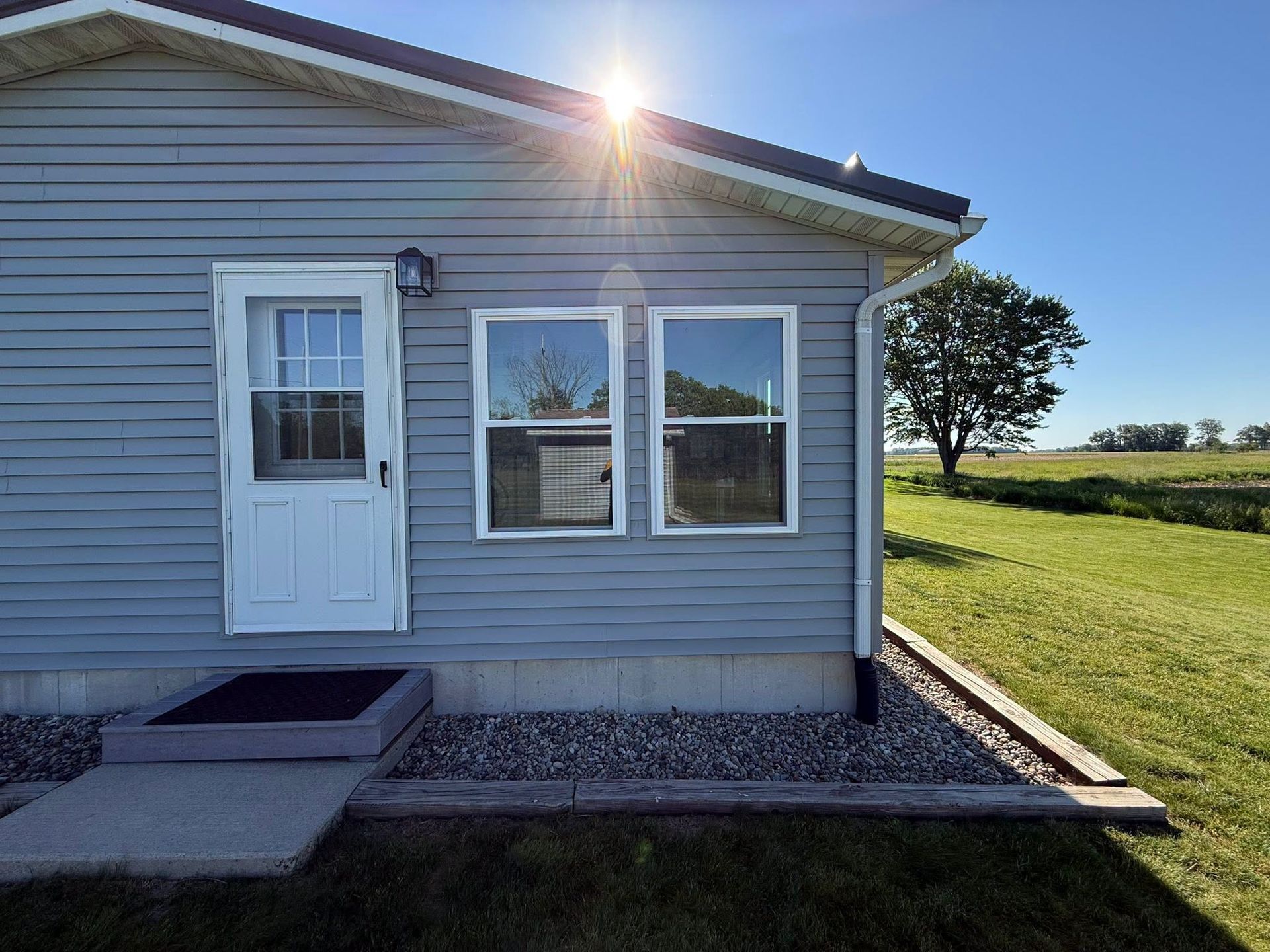Small gray house exterior with white door and windows, front steps, and bright sun over the roof.