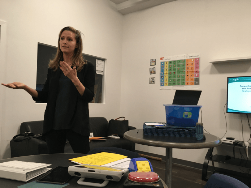 A woman is standing in front of a table in a room giving a presentation.