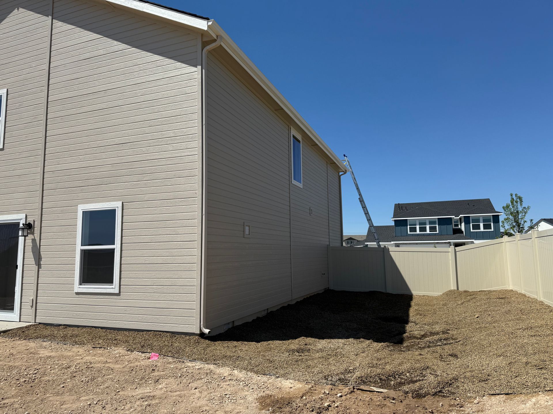 Beige house exterior with a ladder, gravel yard, and a fence on a sunny day.
