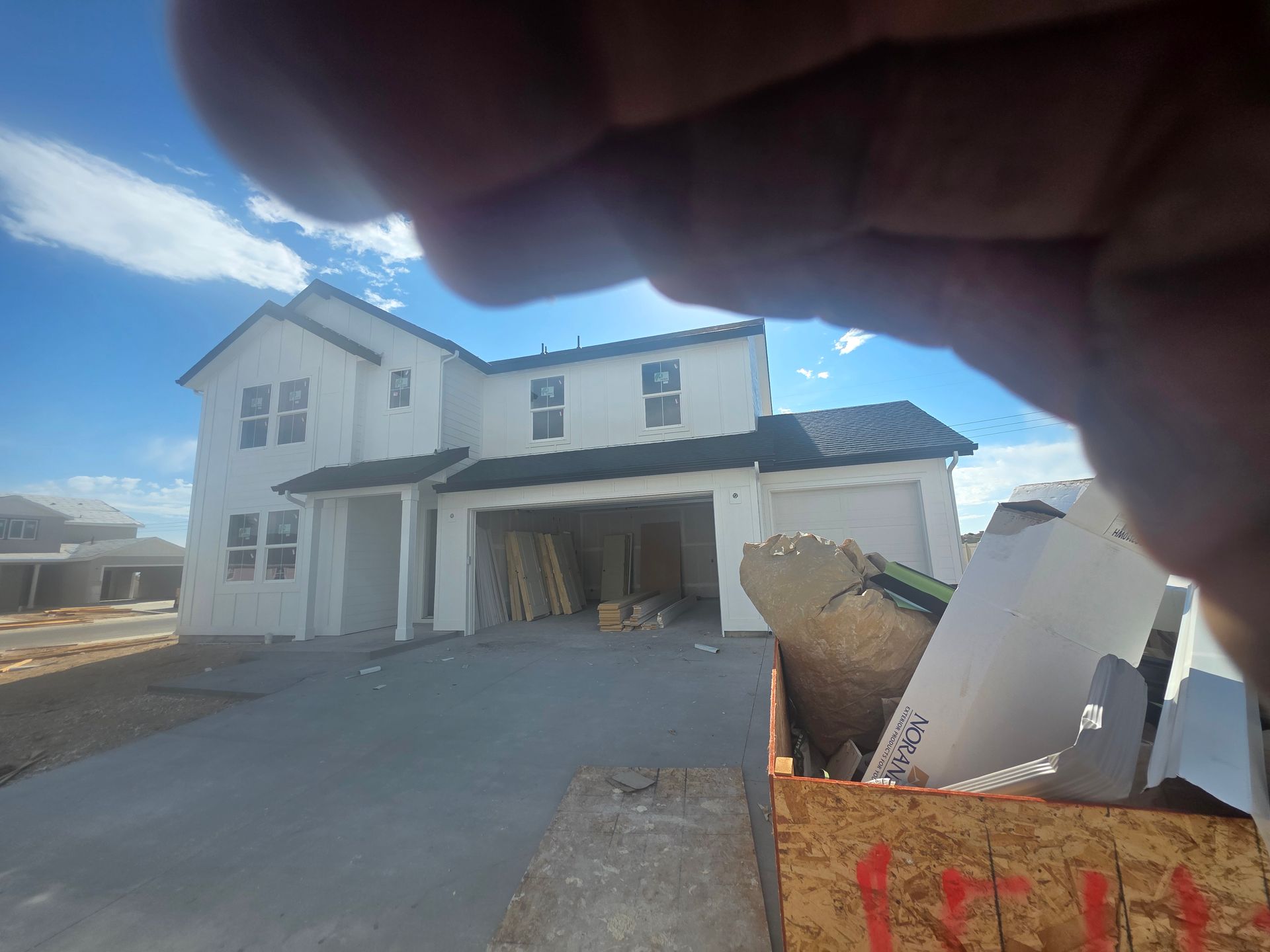 Two-story white house under construction with an open garage, blue sky, and a hand in the foreground.
