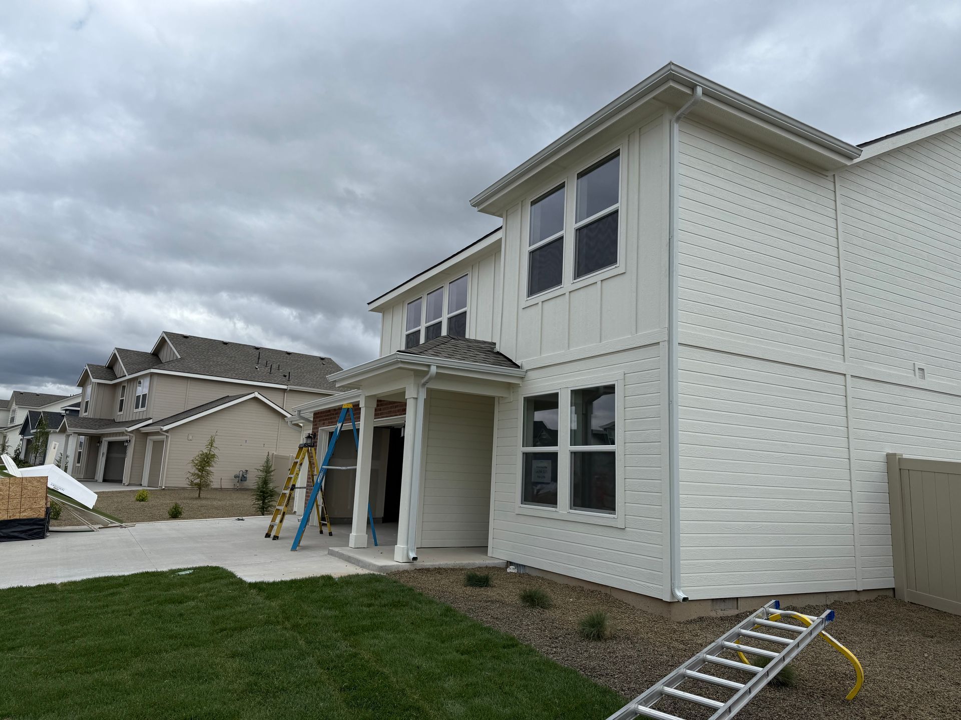White two-story house with dark windows, under a cloudy sky. Lawn in front, houses in the background.