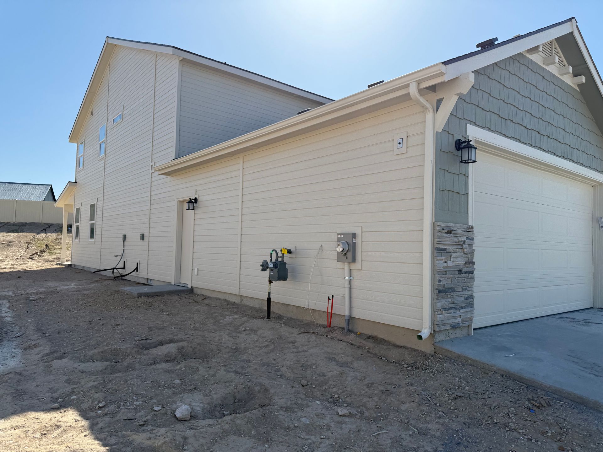 Side view of a two-story house with beige siding, a garage, and a gravel yard under a blue sky.