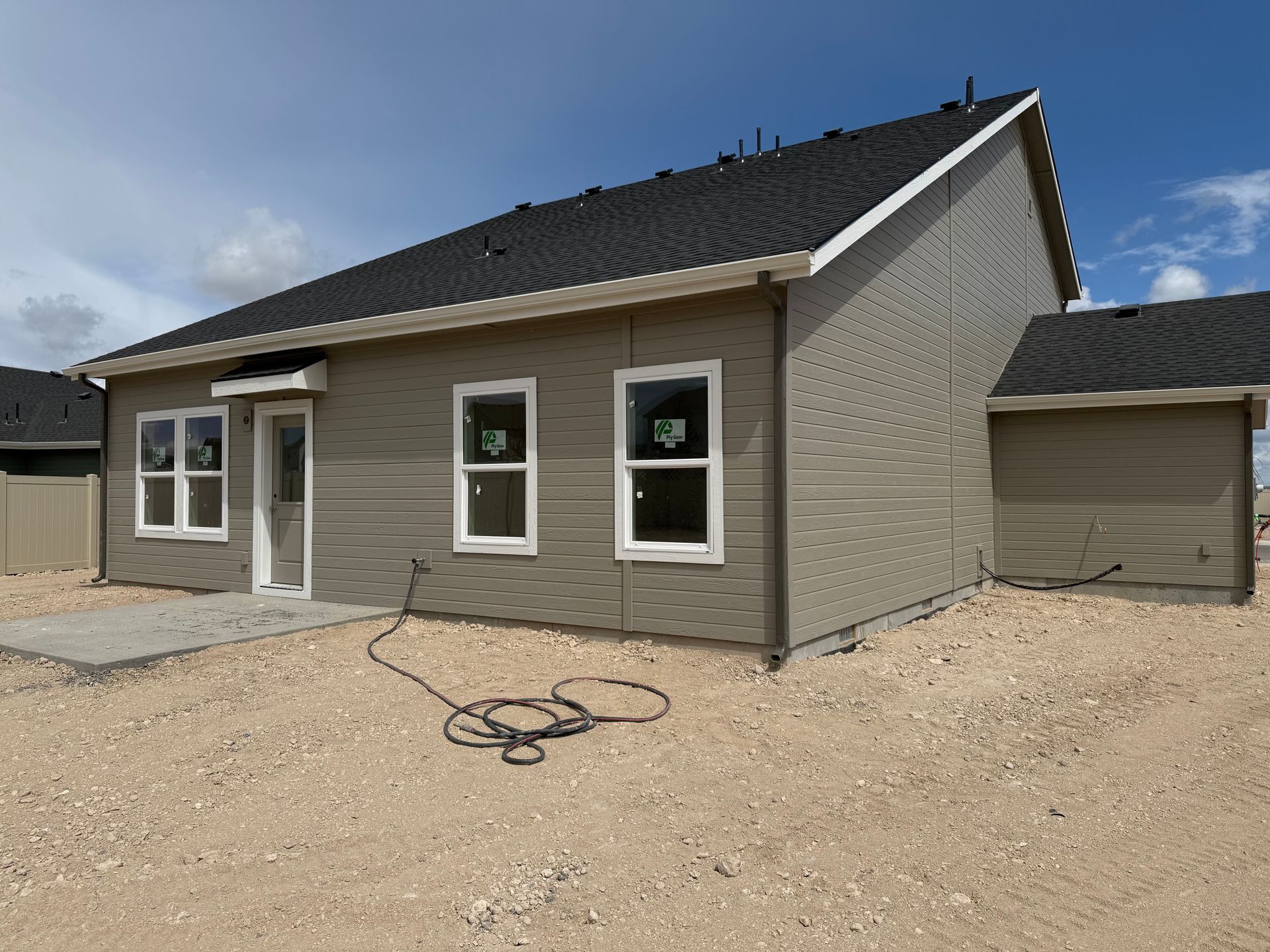 Rear view of a new house with gray siding, windows, and black roof, on a gravel lot under a blue sky.