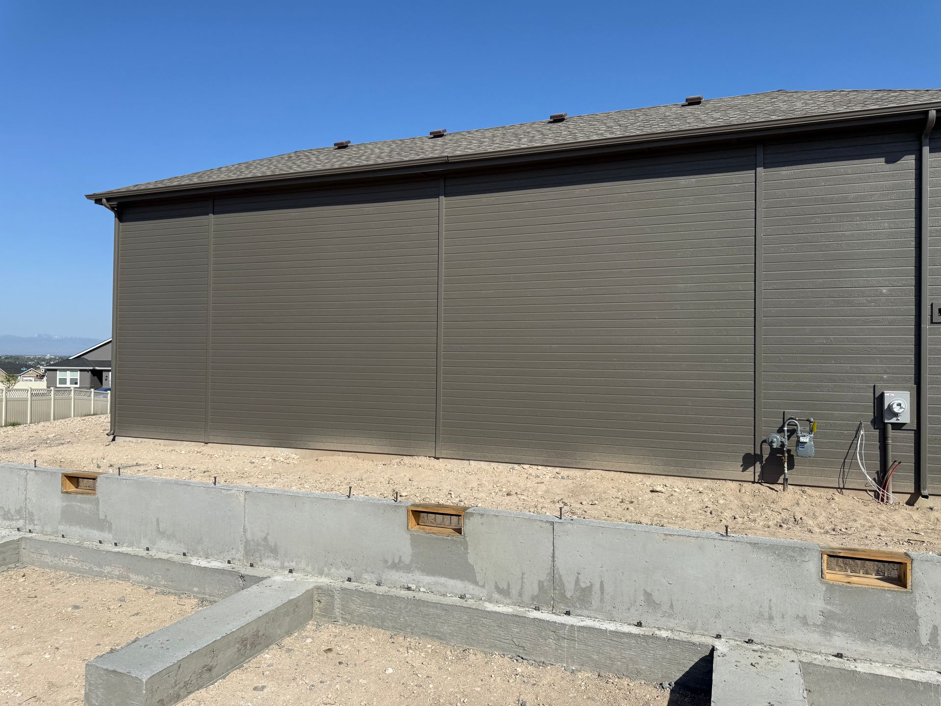 Side of a brown-sided building with gray roof, set on a concrete foundation, surrounded by gravel.