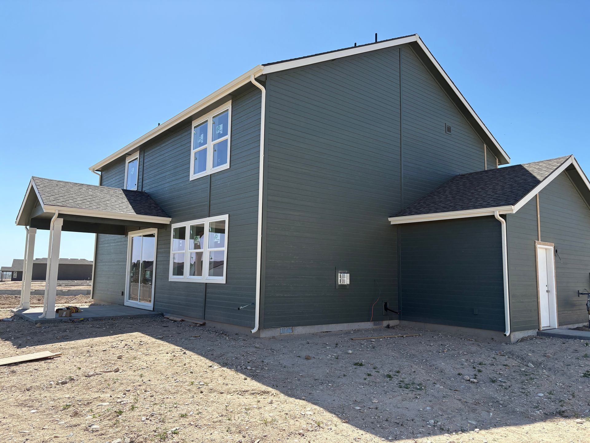 Two-story house with green siding, white trim, and a small porch on a dirt lot under a blue sky.