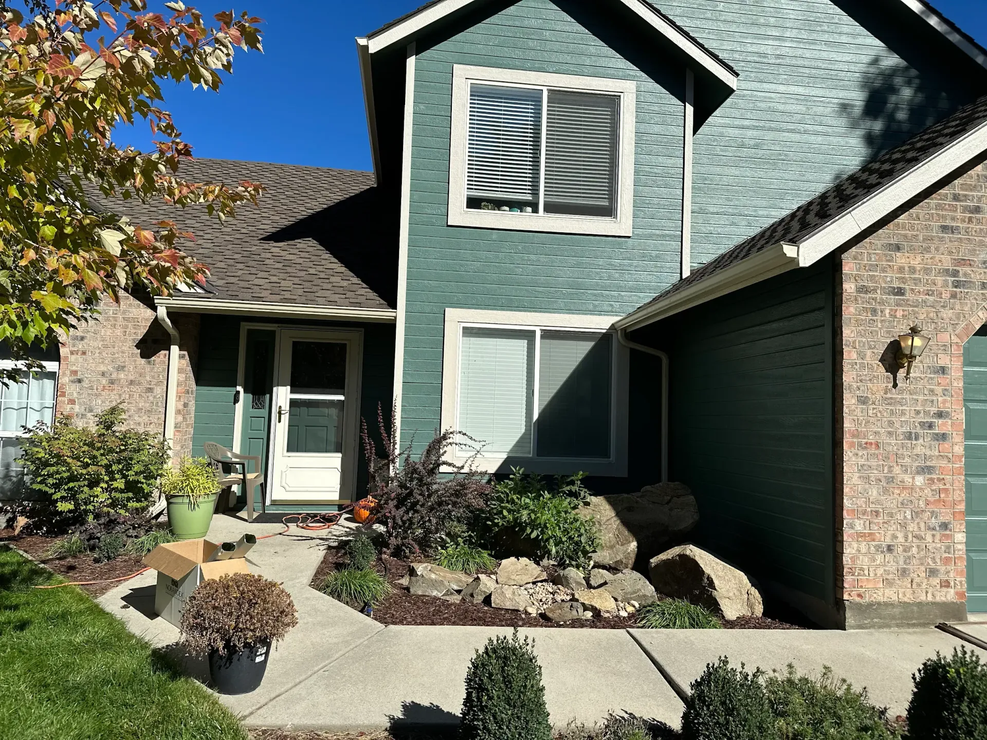 Two-story teal house with brown roof and brick accents. Landscaped front yard with plants and sidewalk.