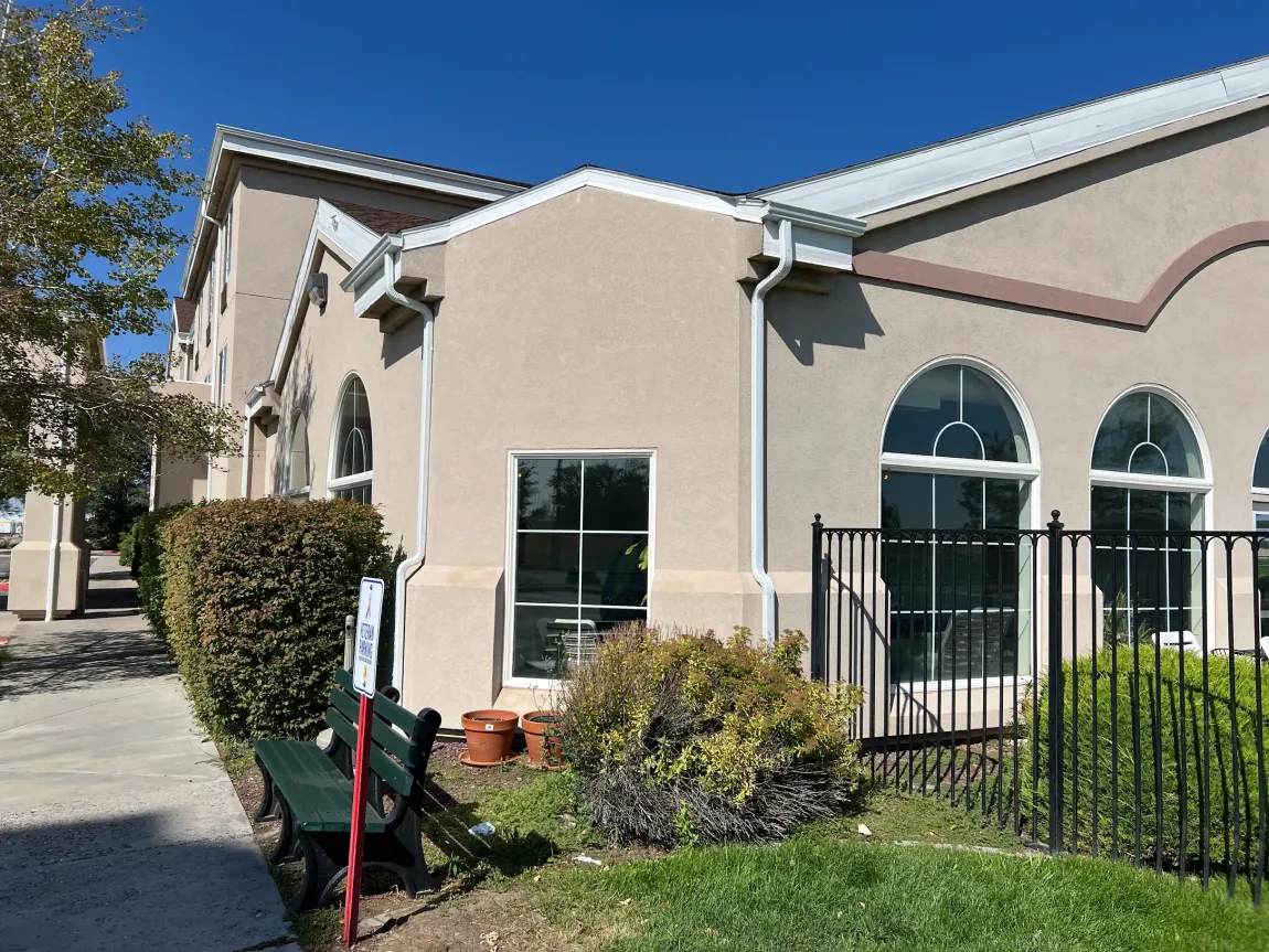Tan building with arched windows, shrubbery, and a bench behind a black fence.