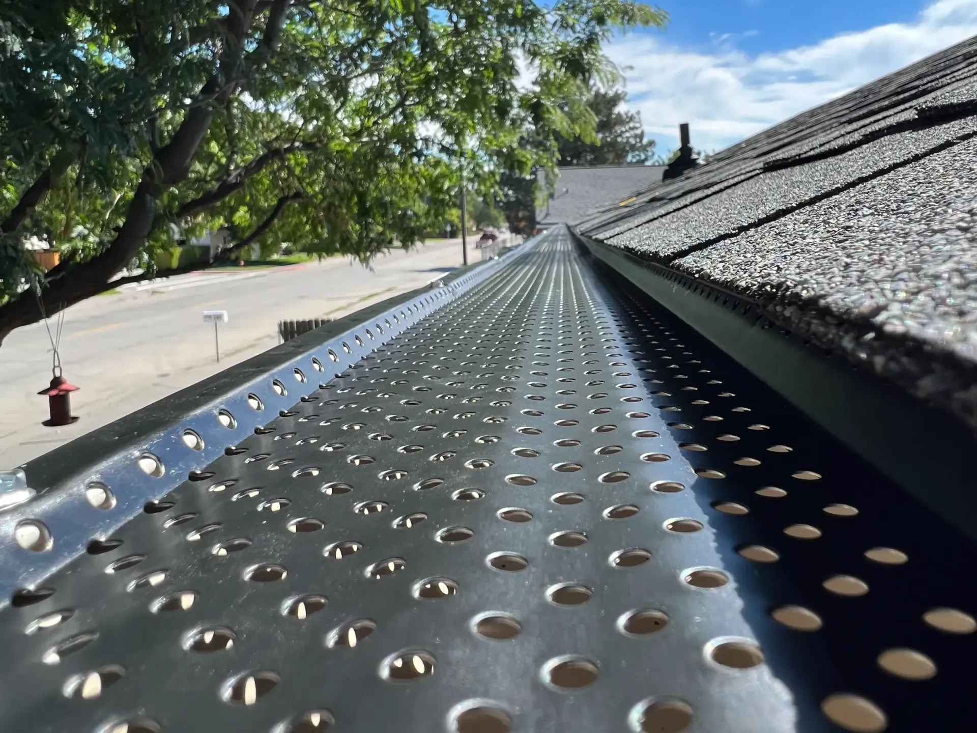 Gutter with perforated metal guard along a roofline, street and trees in background.