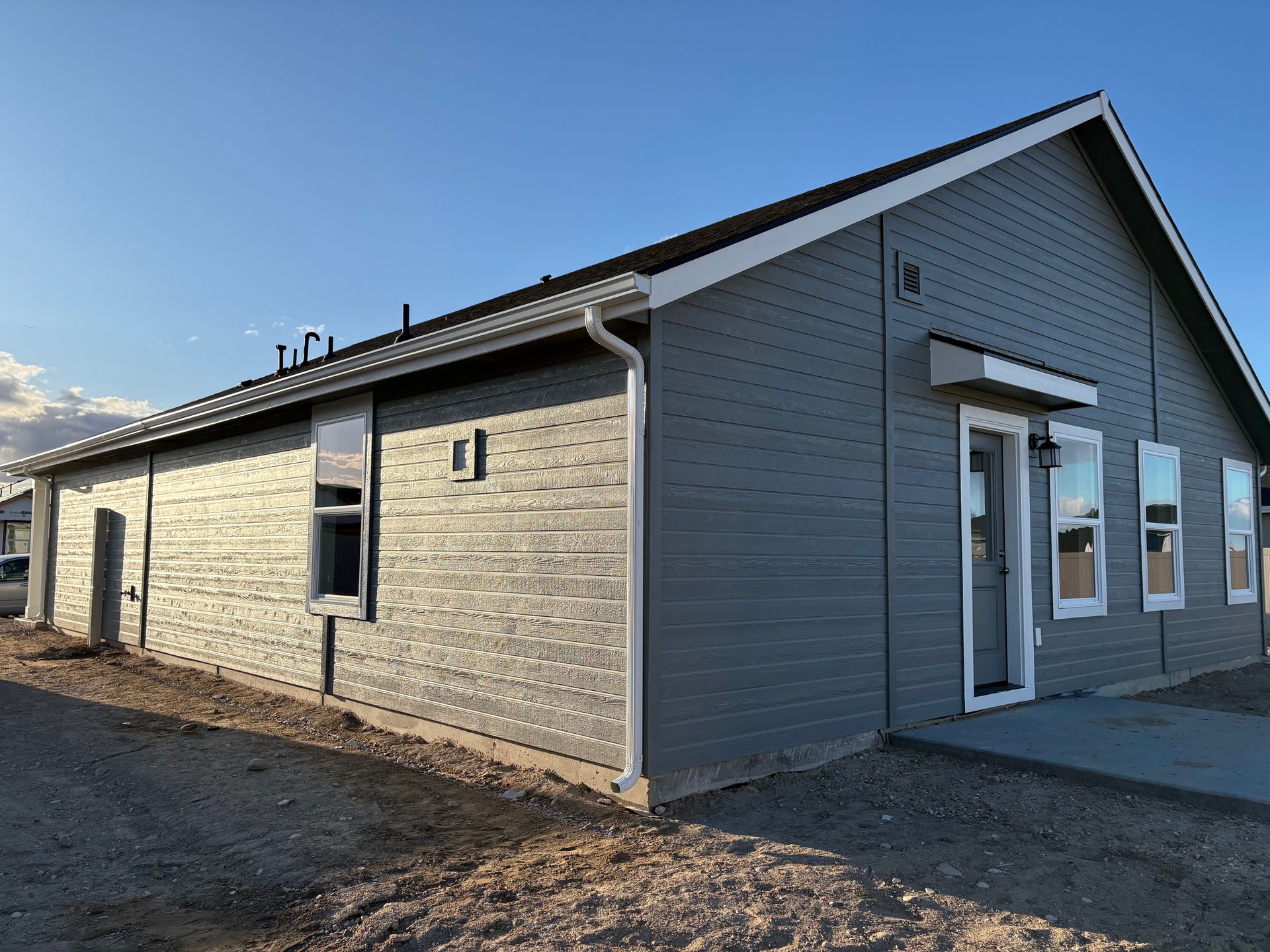 Gray-sided single-story building with white trim, windows, and door, in a gravel lot under a blue sky.