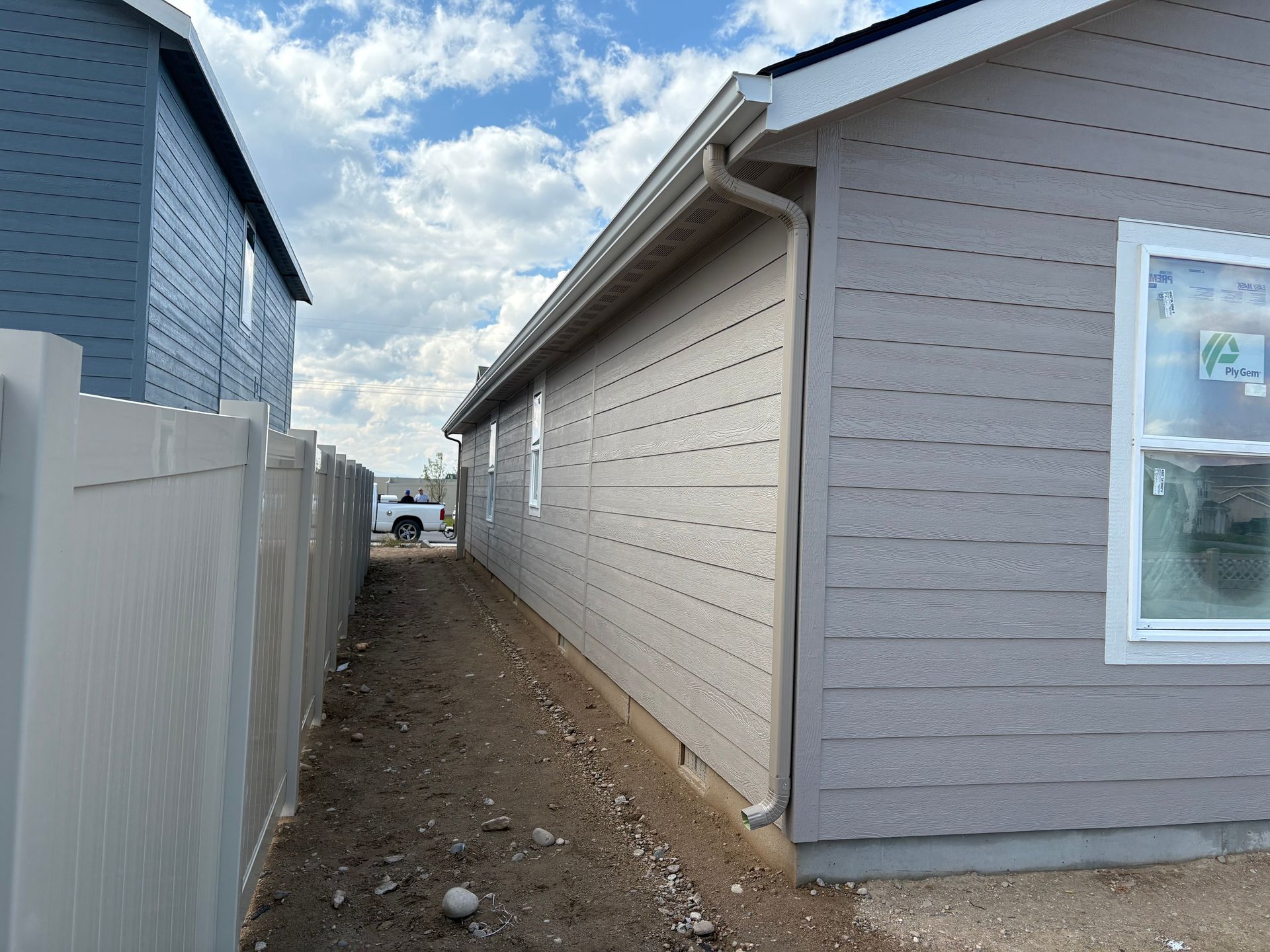 Side view of a house with gray siding next to a beige fence, narrow dirt path in between.