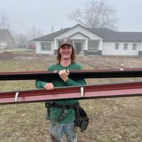 Man holding gutter material in front of a house. He is wearing a green shirt, a hat, and work belt. Cloudy day.