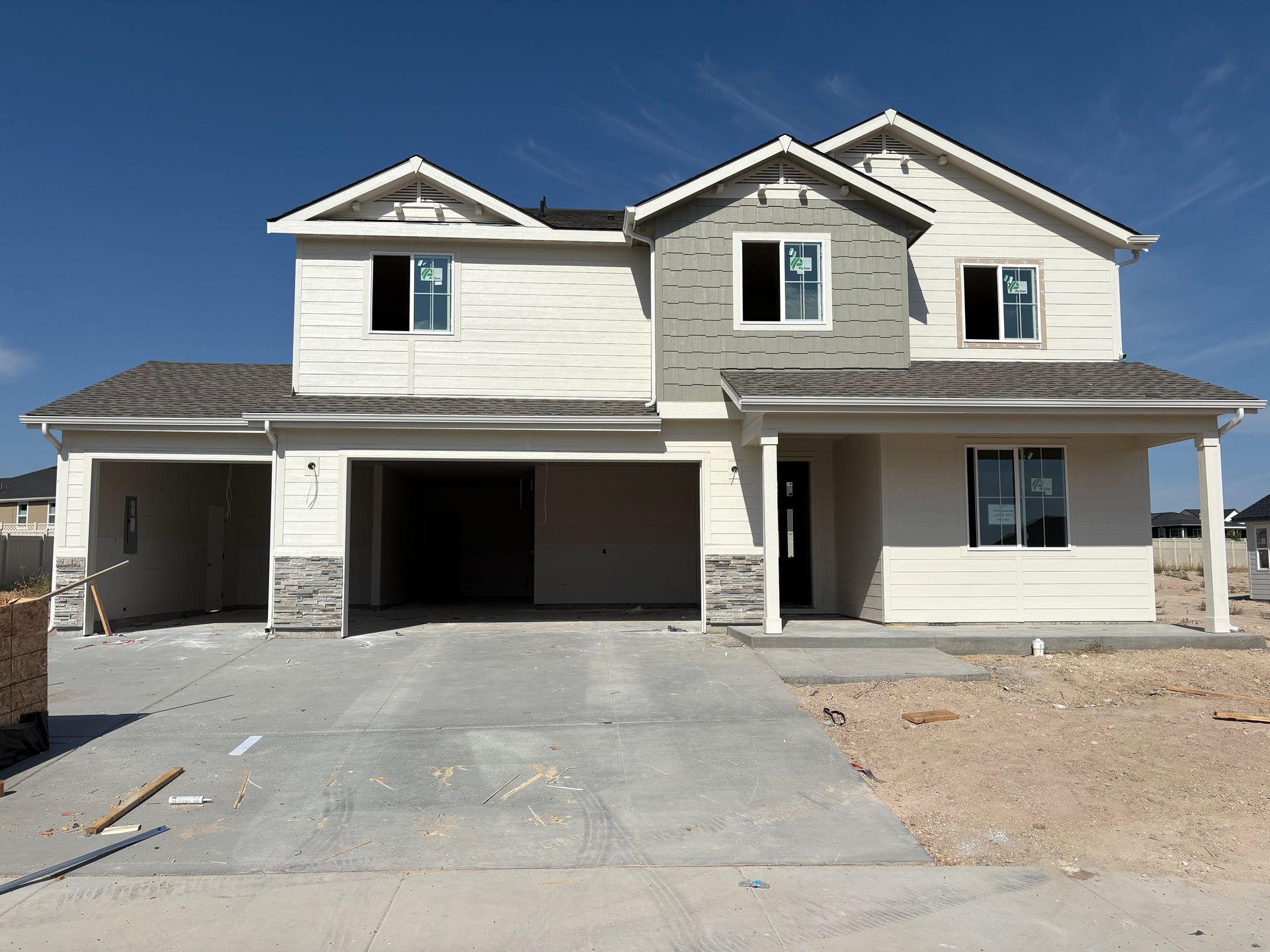 Two-story house under construction with a light-colored exterior and a gray garage door, set against a blue sky.
