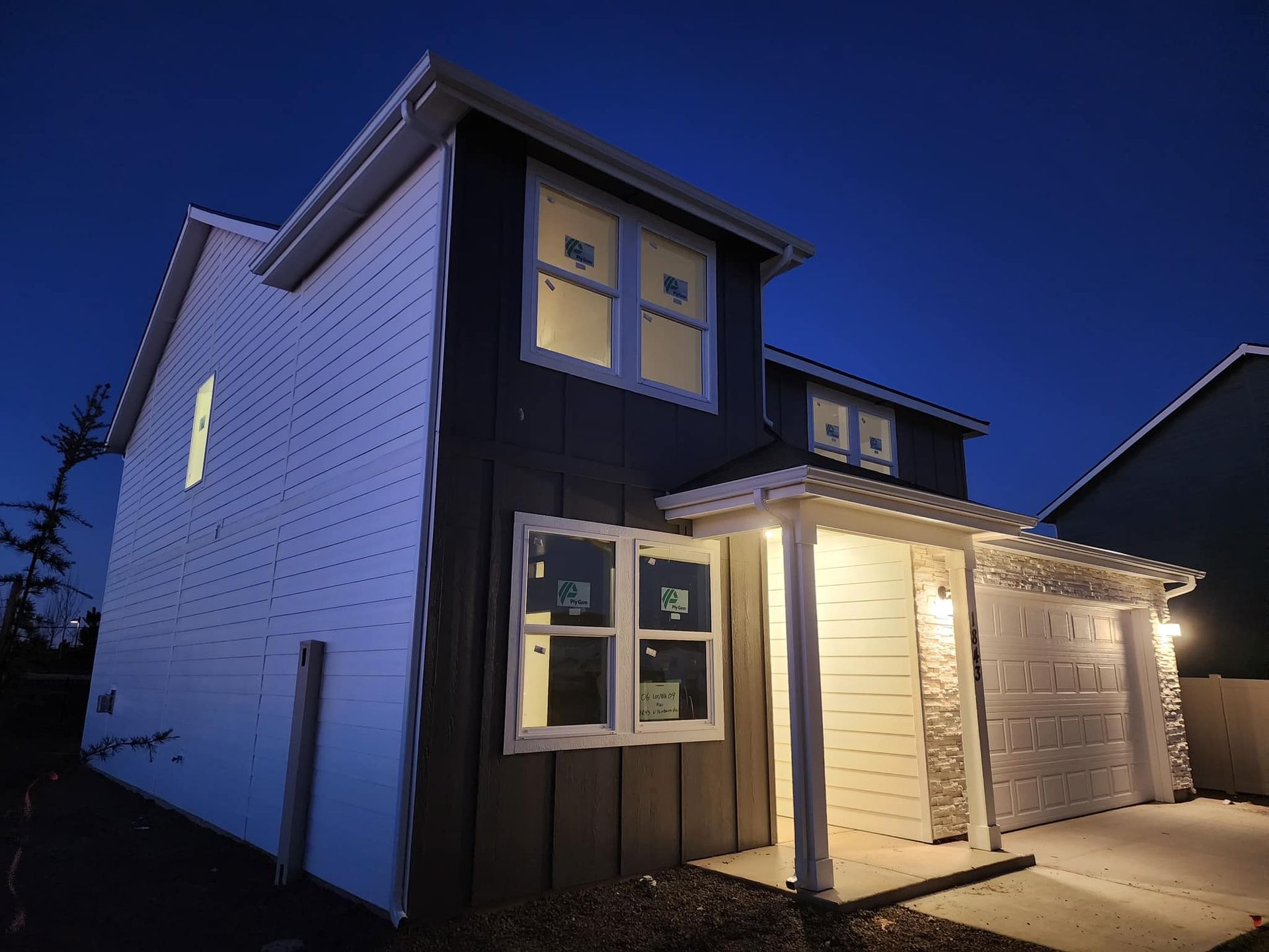 Two-story house at dusk, with lights on inside and outside, gray and white siding, garage door on the right.