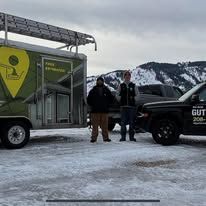 Two people stand by a trailer and truck in snowy conditions. Mountains in background. 