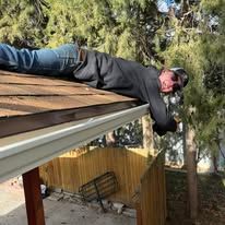 Person lying on a roof, reaching into a gutter; wearing jeans and a black jacket.