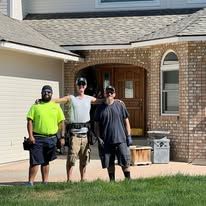 Three people posing near a building's entrance. They wear work gear; green, grey, and black shirts.
