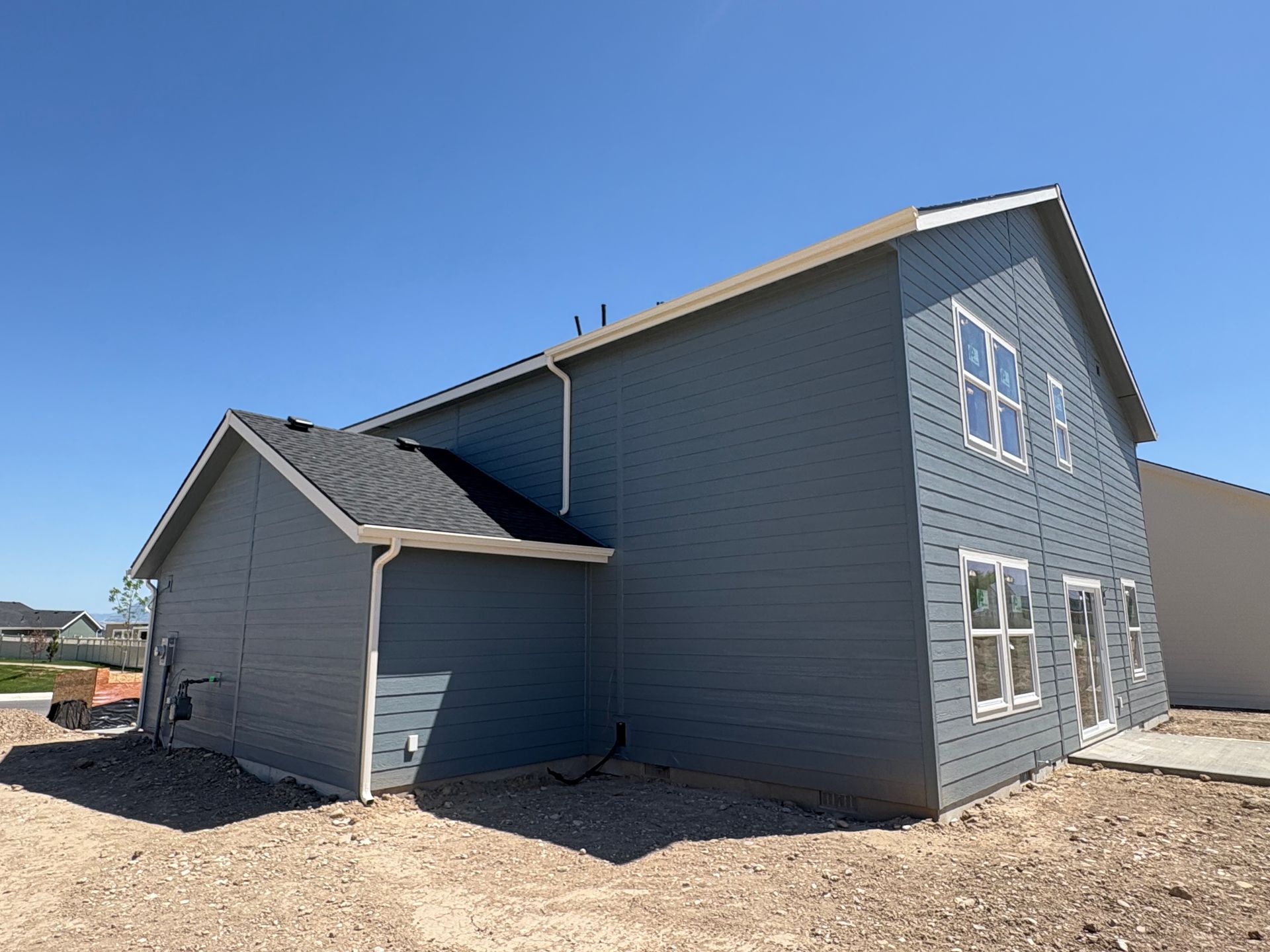 Blue-sided house with white window frames and brown trim on a gravel lot under a clear blue sky.